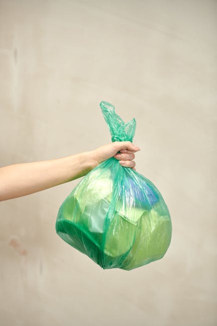 A person’s hand is shown holding a green plastic rubbish bag tightly by the tied top, with the bag appearing full and slightly bulging. The bag is made of translucent plastic, allowing some contents