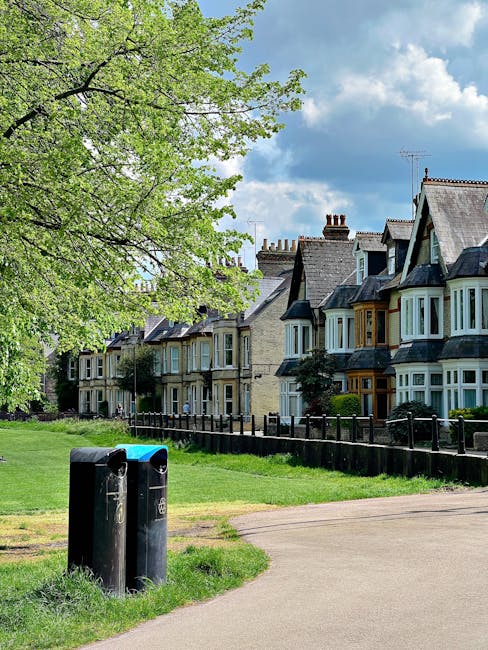 A pair of black cylindrical rubbish bins with blue and green lids are situated on a grassy verge alongside a paved footpath in a residential area. Behind the bins, there is a row of Victorian-style te